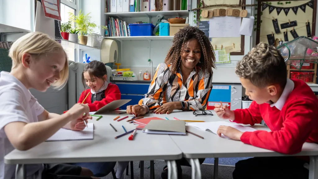 A classroom scene showing a group of children seated around a table with notebooks and coloured pencils, engaged in an activity. A teacher sits with them. The background includes shelves with books, storage boxes, and educational materials, along with a display board and various classroom decorations.