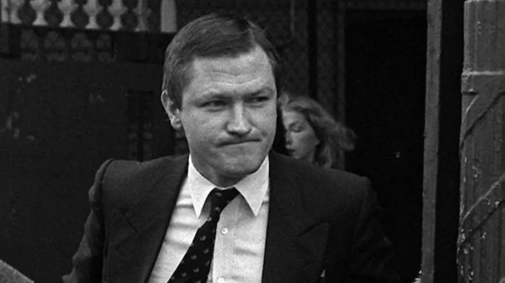 Black and white image of Pat Finucane. He's wearing a with a tie. His lips are pressed together. He has come out of a gate. A woman is also coming out of the gate.