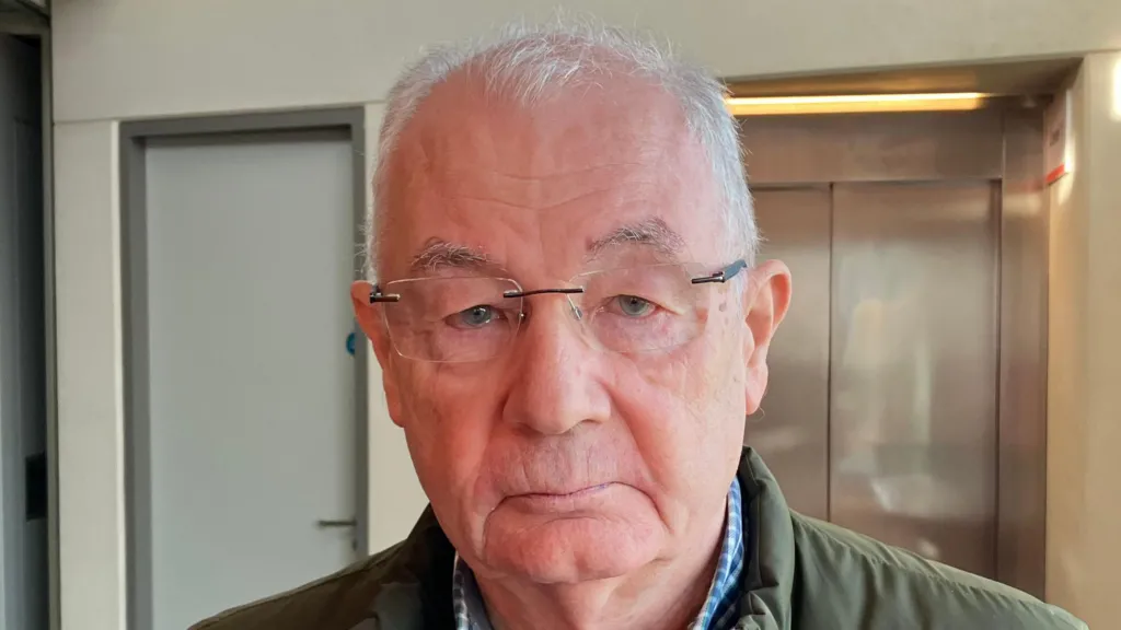 Mickey McKinney has short grey hair. He is wearing glasses and has a green coat on with a blue shirt and jumper. He is standing in a reception area with a lift in the background. 