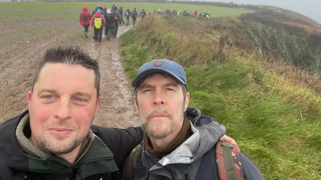 Craig Maxwell smiles and has an arm around comedian Rhod Gilbert's shoulder. The pair are in the middle of a muddy path with a group of hikers in the background.