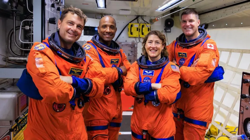Four smiling astronauts wearing bright orange spacesuits but no helmets. From left to right they are Reid Wiseman, Victor Glover, Christina Koch and Jeremy Hansen. They are standing together positioned in a row with their arms crossed. The room they are in is filled with technical equipment and cabling.