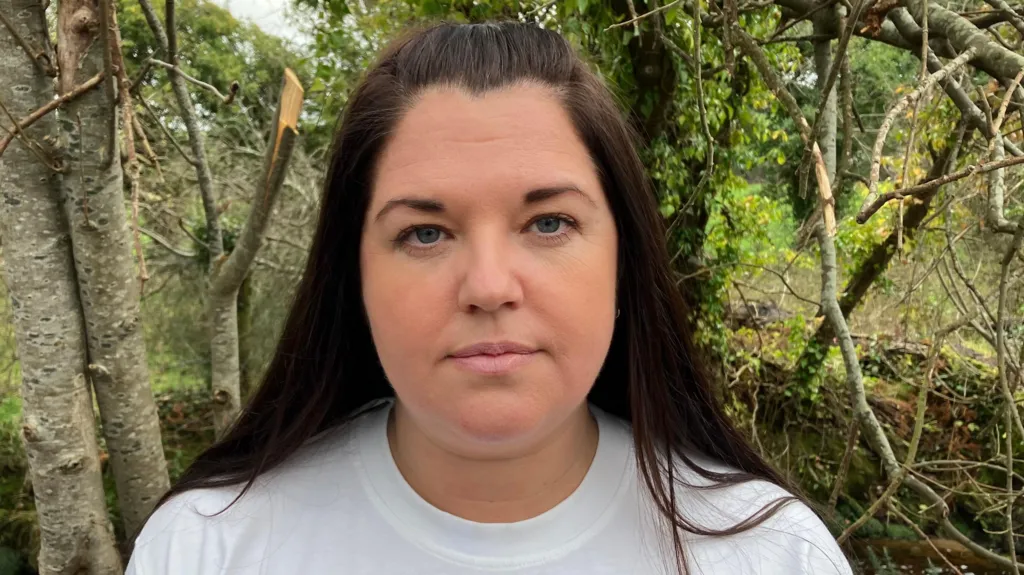 Gail has a white T-shirt on and long brown hair, some of which is tied back. She is standing in front of some trees.