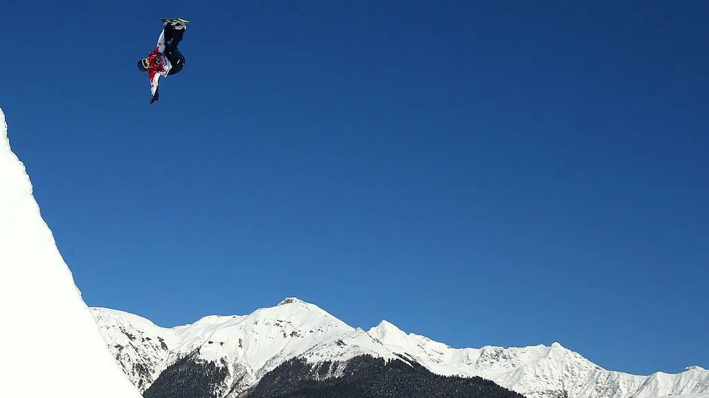 Aimee Fuller in a red and white coat mid jump with blue sky and mountain tops in the background 