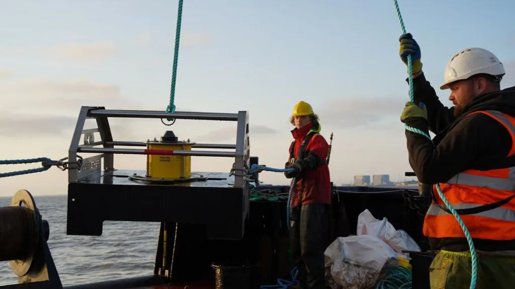 Two people in hard hats and high vis jackets stood near water with machinery they are pulling out of it. 