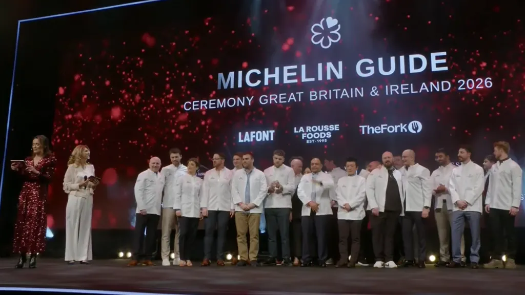 A group of chefs standing on stage in white jackets during the Michelin Guide Great Britain & Ireland 2026 awards ceremony in Dublin, with presenters at the side and a large illuminated backdrop displaying event branding. Danny Young is the chef on the left and and Tom Tsappis is sixth from the left. 