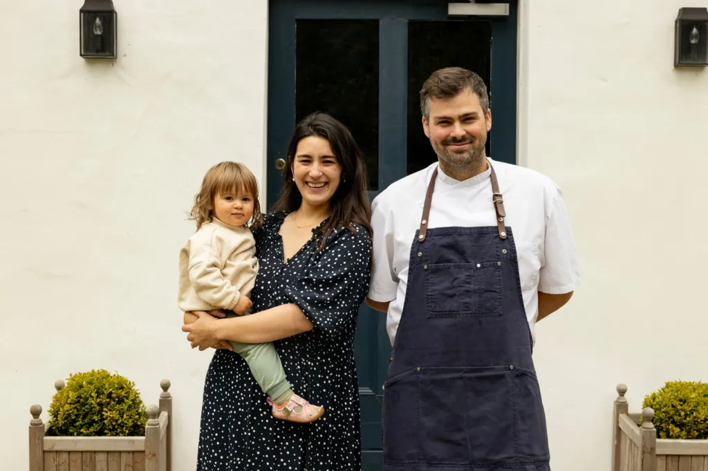 Matilda and Tom Tsappis standing outside Killiecrankie House. Matilda has shoulder length dark brown hair and is wearing a black dress with white spots and is holding a toddler on her hip. Tom has short brown hair and a trimmed beard. He is wearing a white chef's jacket and a navy blue apron with leather neck strap.