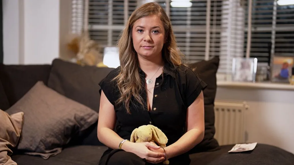 Chloe, in a black button‑up shirt sits on a dark couch, gently holding a yellow stuffed bunny. Behind her are cushions, a window with blinds, and framed photos on the sill.