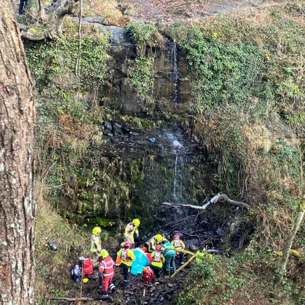 An outdoor scene of rocks and greenery shows a ledge at the top with a small waterfall trickling off.  At the bottom of the photo, a group of mountain rescuers and firefighterss are huddled around someone who has been covered by a blanket.