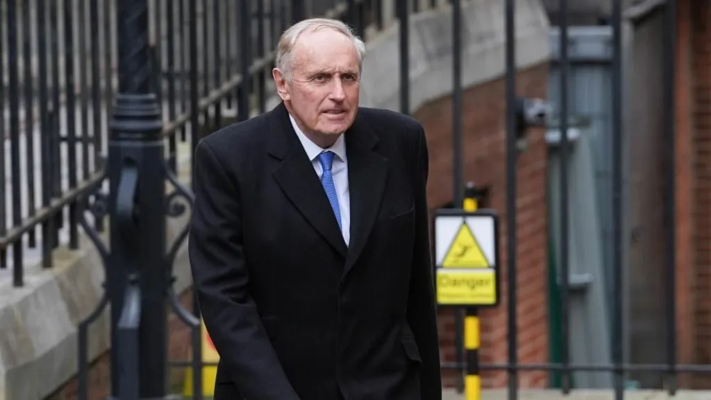 Dacre walking in front of a black metal fence. He wears a long black coat, a white shirt and blue tie. He looks sincere 