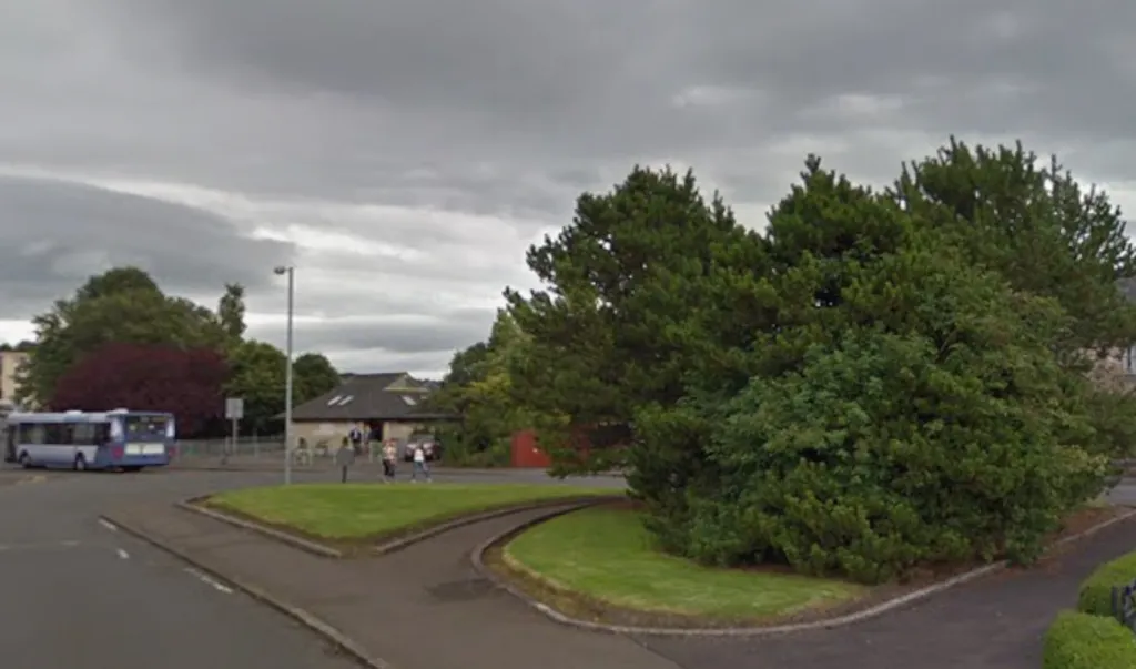 A footpath with a large tree in the middle. A road and bus are visible in the background