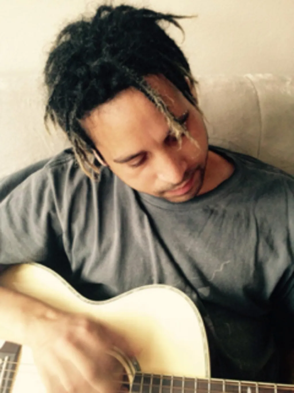Joe Black sits, strumming his guitar. The picture is taken from a perspective looking down on Joe from above so you can see his dark, dreadlocked hair, highlighted blond at the tips. His eyes are partially closed and his lips together as he plays. You can see a short black stubble goatee. Joe identified as mixed white and black Caribbean/Jewish, according to his family, and he also had Irish heritage.