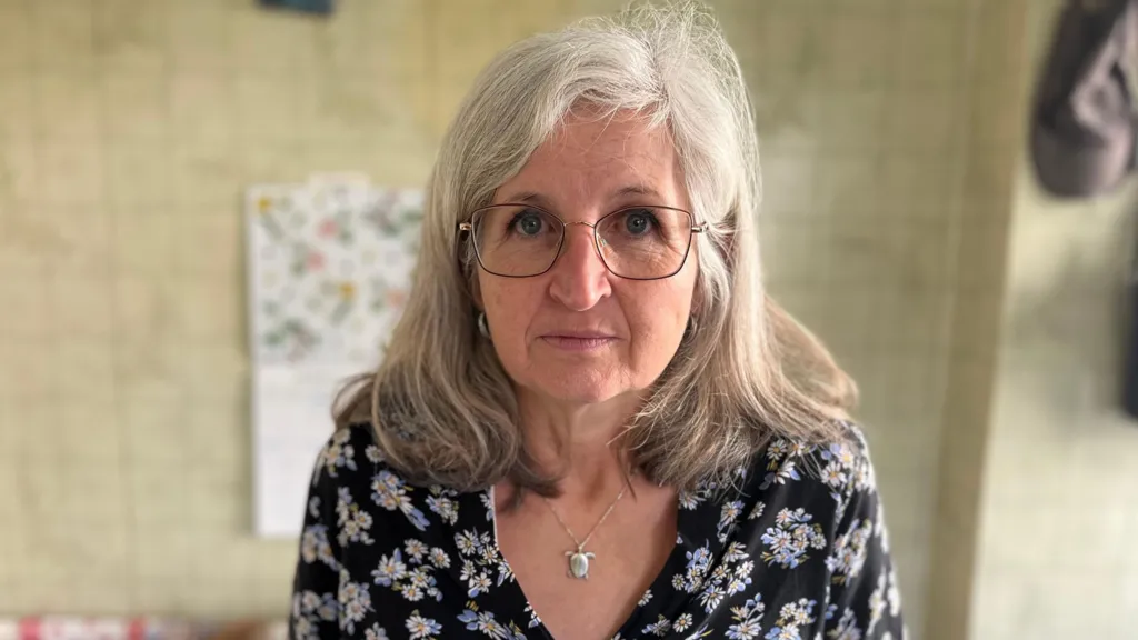 A woman with grey hair, glasses and a dark blue blouse with flowers is looking into the camera. She is standing in her kitchen. 