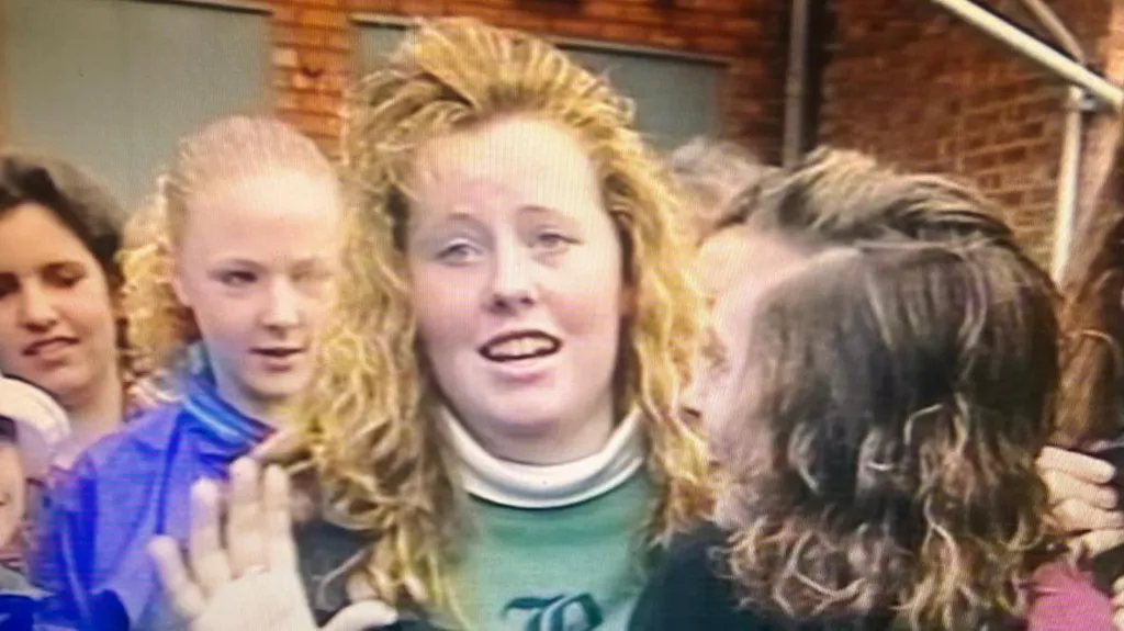 A still from footage filmed in the 90s of Jemma Williamson as a teenager. She has blonde hair and a green jumper. She is in the process of talking and is in a crowd of young woman