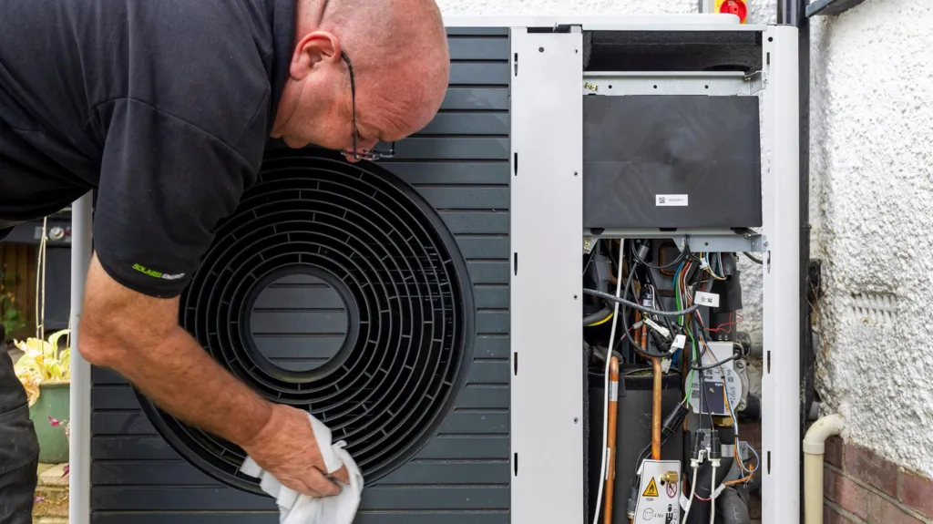 An engineer - a man wearing a black T-shirt - wipes the outside of a black heat pump with a cloth. To the right of the picture the heat pump abuts the wall of a property with the pipes and electrical components visible