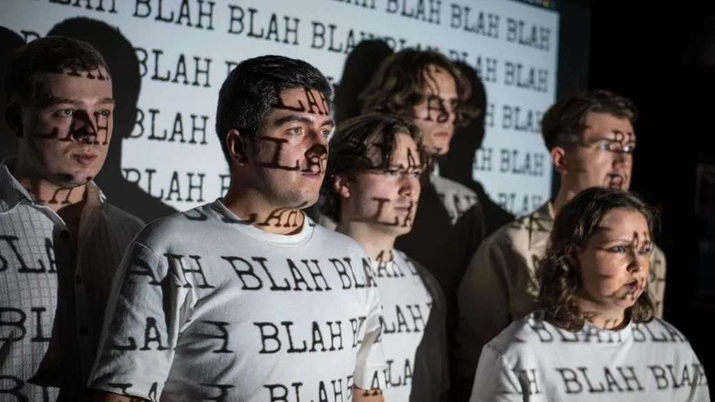 A group of six young people on stage, looking to the right. They are wearing white T-shirts and standing in front of a white screen. The word BLAH in black capitals is being projected onto them and the screen. They look very serious.