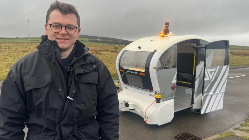 A man with dark hair and glasses in a big black jacket stands in front of a white driverless vehicle on the road with its door open