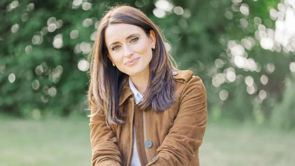 A glamorous image of Anne taken at the Royal Highland Show - she wears a tan suede jacket and her hair is long. She is smiling and there are trees and grass behind her.