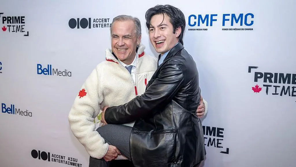 Prime Minister Mark Carney, wearing a beige fleece sweater with a red Canadian maple leaf on the arm, laughs as he hugs Heated Rivalry actor Hudson Williams, wearing a leather jacket.