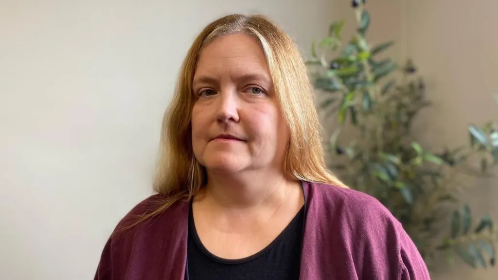Anna has long blonde hair. She is wearing a black top and a purple cardigan. Behind her are the green leaves of an indoor plant against a white wall.
