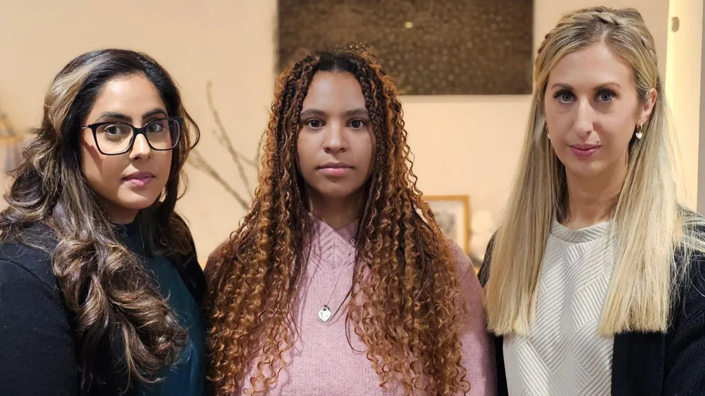 Three women stand together, looking straight at the camera. One wears glasses and a navy blue top, one has a pink top and the third has a white top under a dark cardigan. All have long hair.