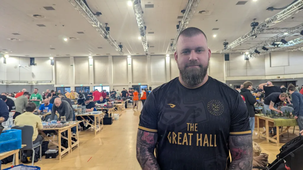 Liam Dempsey, a man with short hair and a beard, is stood in the hall. He's wearing a black sports shirt and has two sleeves of tattoos. 