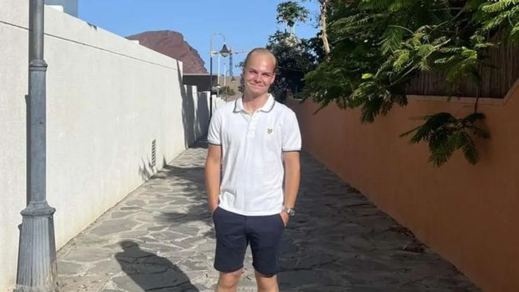 Ellis Pears standing in an alleyway with large paved stones on the floor. He is wearing a white polo t shirt and blue shorts with his hands in his pockets. There are trees encroaching on the alleyway, which has an orange wall, from a garden to  the right of the image.