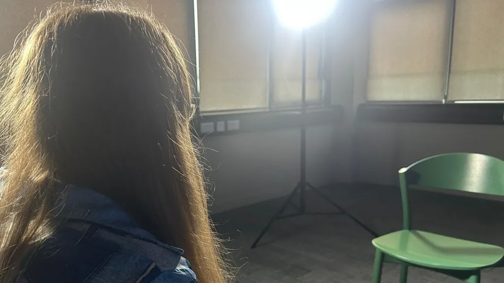 Susan is sitting to the left of the picture but you can only see her shoulders and her hair. In front of her is a green chair, and there is a large light in the corner. She is in an empty room with screens pulled down 