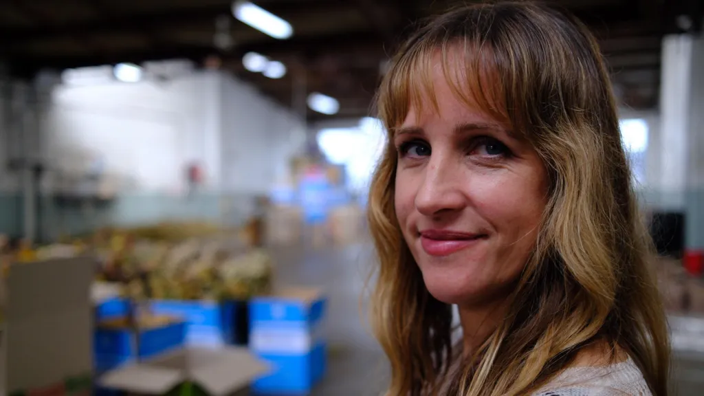 A blonde woman smiles at the camera, boxes of flowers in the background
