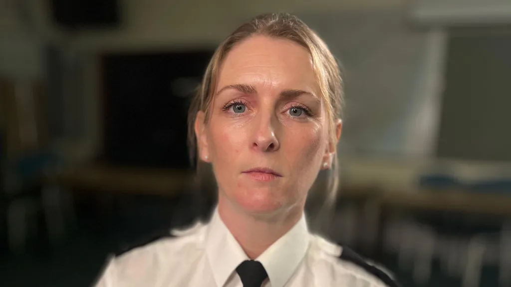 Assistant Chief Constable Alice Scott looks at the camera expressionless. She has blonde hair that has been tied back behind her head, with some pieces left out that frame her face. She wears a white shirt with a black tie and black lapels. 