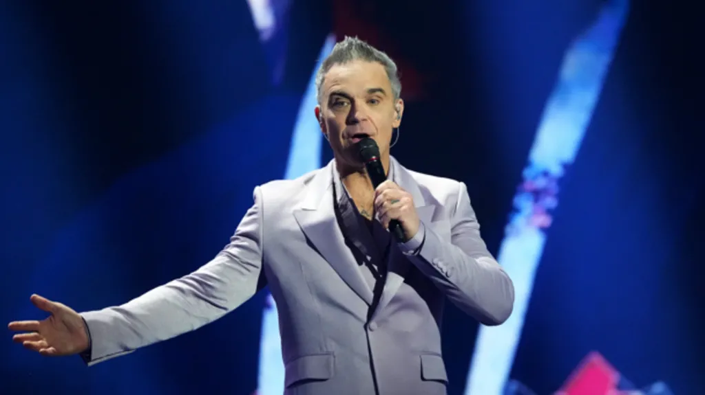 Robbie Williams, who has grey hair and is wearing a grey suit with an open collared shirt, stands on stage holding a microphone in his left hand
