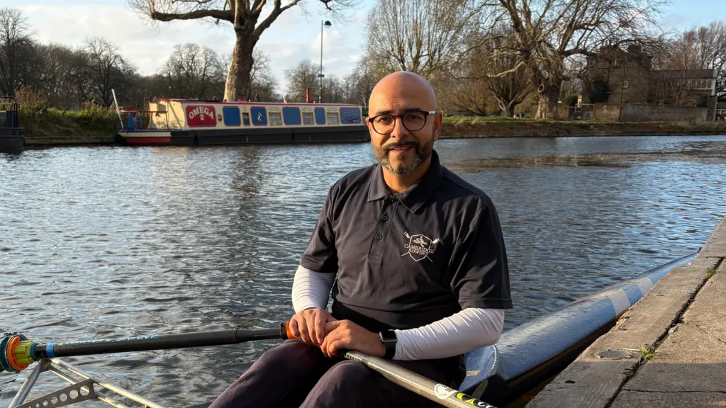 Omar sitting in a single scull rowing boat on the side of a river. He is smiling slightly, while holding an oar in each hand. It is a fine, bright day. He is wearing a dark blue, collared T-shirt, with all three buttons done up, though his arms are covered by a white top worn underneath the T-shirt. He is wearing dark blue jogging bottoms and the tops of his knees are just in shot. His boat is against a pontoon edge, which is to the right of frame. Behind Omar is the far river bank, with a narrow boat moored up, a grassy bank, trees and a building. The river looks calm and it is a bright day.