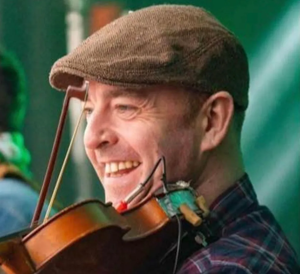 A man smiling and wearing a brown flat cap and red, white and dark blue flannel top. He has a violin under his chin