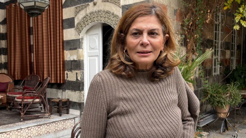 Kabawat in the garden of her home, which is a traditional Syrian house built in lines of light and dark brick, with an ornate arch over a white wooden door, decorative tiles on the steps and climbing plants around the windows. She wears a light brown jumper and is looking a little to the left of the camera with a thoughtful expression.