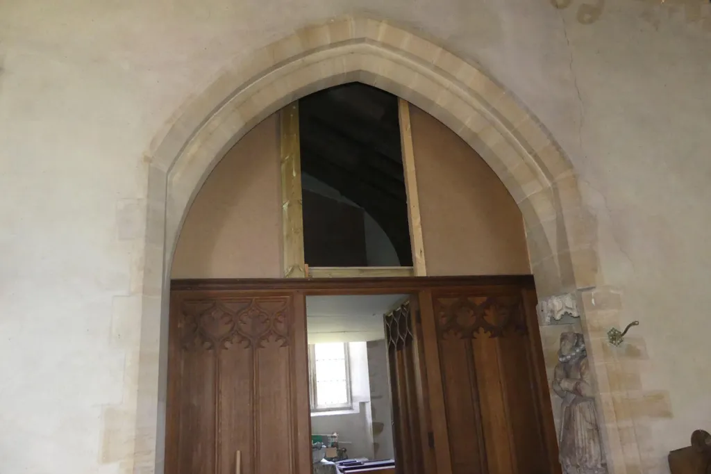 Inside a church, a brown ornate door is open and the vestry can be seen through it. Above it, another door is open to show the bat loft. It is dark inside, but the rafters of the roof can just be made out.   The wall surrounding the open doors is pale grey stone, with an ornate carving on the right hand side.