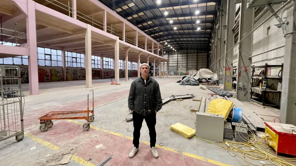 A man in a black jacket, black trousers, white trainers and a white woolly hat stands in a huge foundry. There are hardware trollies, tools, building materials and equipment covered by tarpaulin. To the left, a newly built mezzanine structure. The huge building is well lit and covered by a new roof. There is graffiti on the far wall beneath the mezzanine.