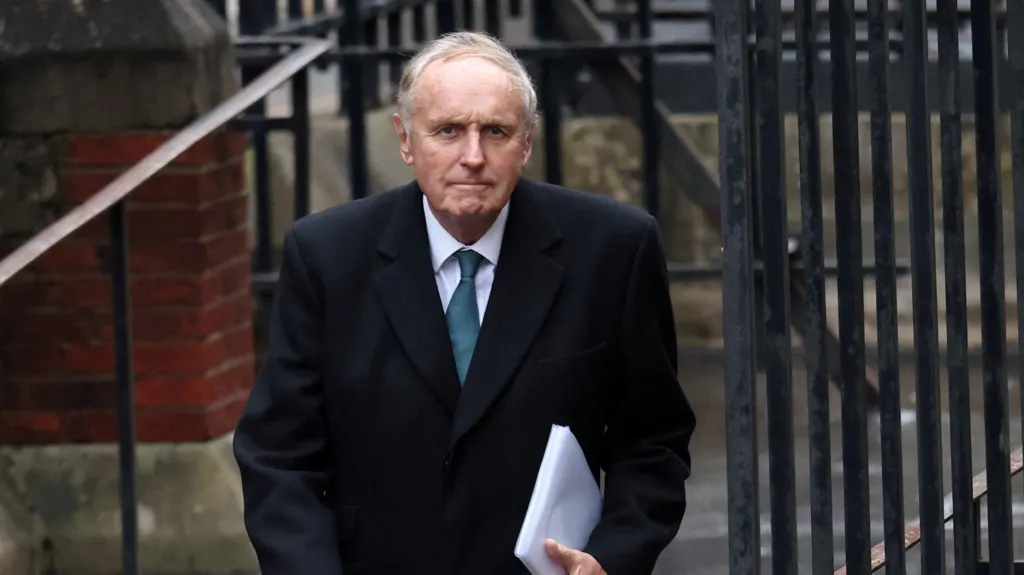 Paul Dacre wearing a formal black coat, white shirt and turquoise tie. He is carrying a wad of white documents under his left arm. he is outdoors. Behind him is black railing and a brick wall of a building