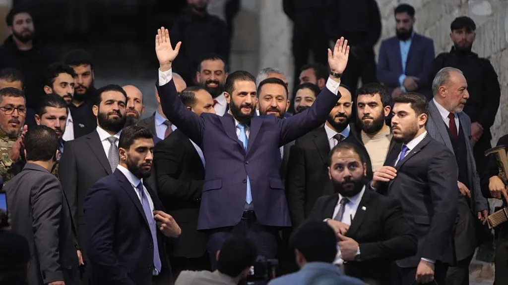 President Ahmed al-Sharaa, wearing a navy suit and blue tie, raising his hands in the air, at an event in November 2025 to celebrate the anniversary of the end of the Assad regime's control of Aleppo. He has dark hair and a dark beard, and is surrounded by other men in suits.