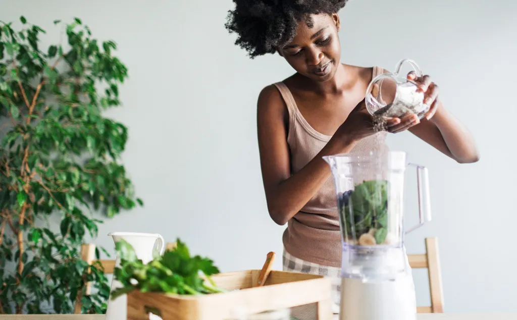Stock photo shows a woman adding chia seends into a smoothie at home, there is a plant in the background.