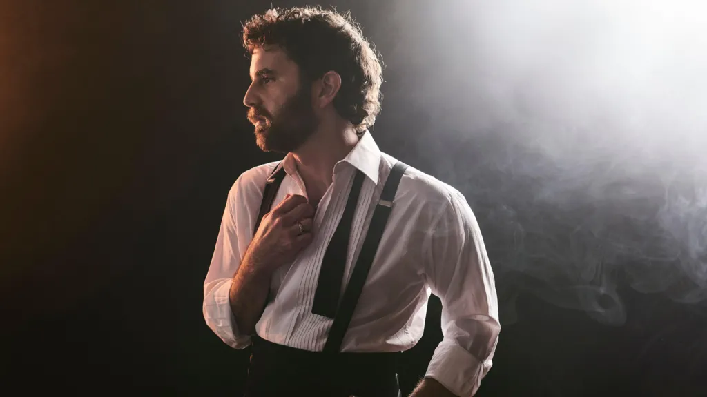 Ben Platt stands under dramatic theatre lighting. He's wearing a smart, white shirt. It's open at the collar, he's wearing braces and a black tie lies untied around his neck. He is looking to his side, into the distance. He has dark hair and a dark beard and lighting is shining from above his left shoulder.
