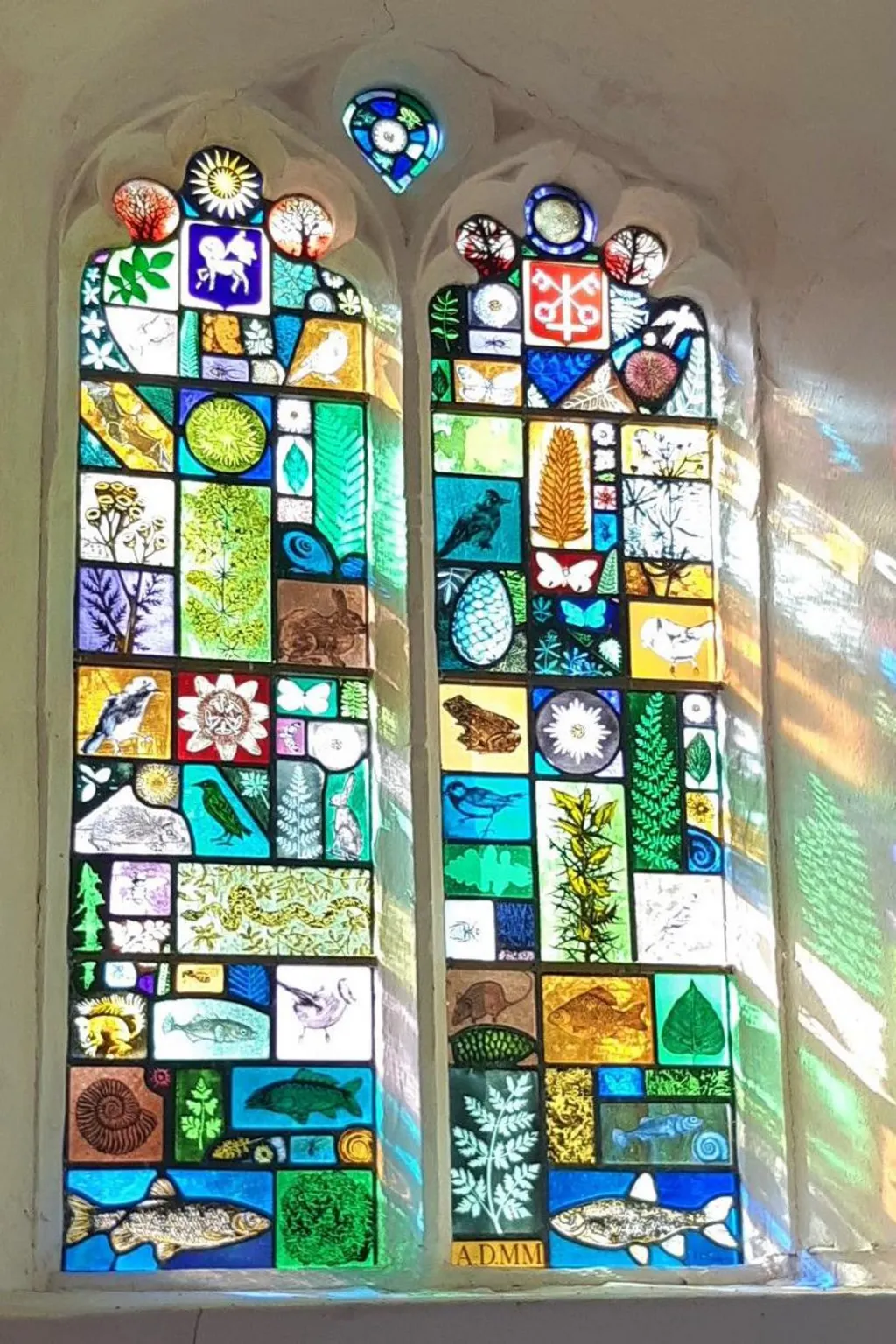 Stained glass window inside Guestwick church. The sunshine beams through it, highlighting colourful stained glass images of nature, including a bat, fish, birds, and leaves from plants and trees. 