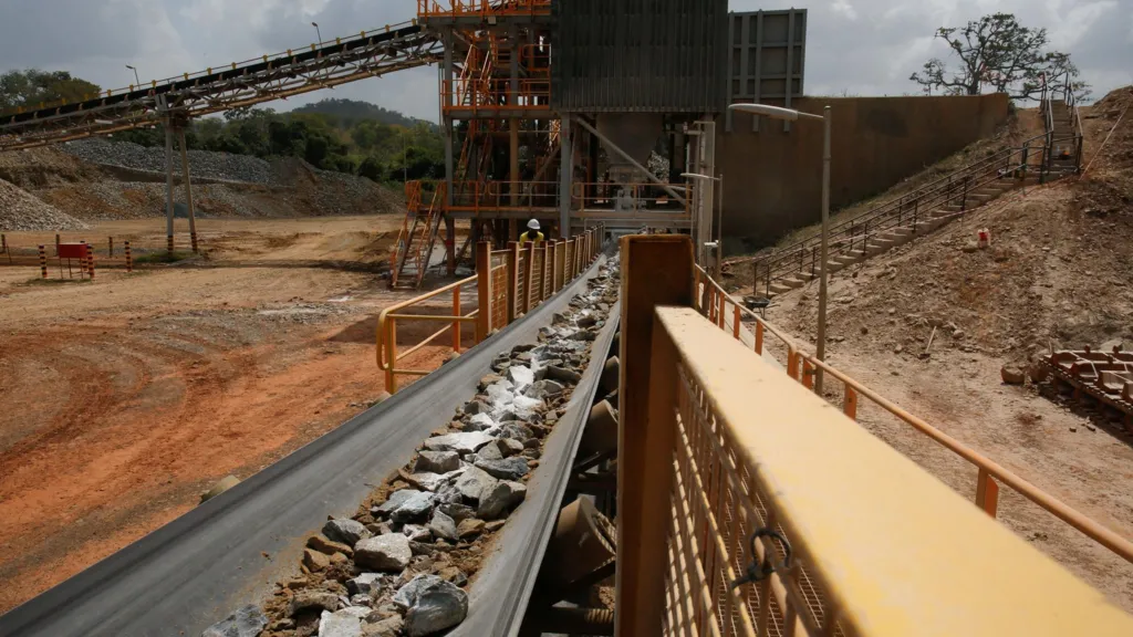 A conveyor belt carried a mixture of rocks and fine particles to a central building in the background. There is a yellow barrier to the right of the belt. Two workers in the background sort through the rocks. The surroundings are orange dusty ground with a few trees