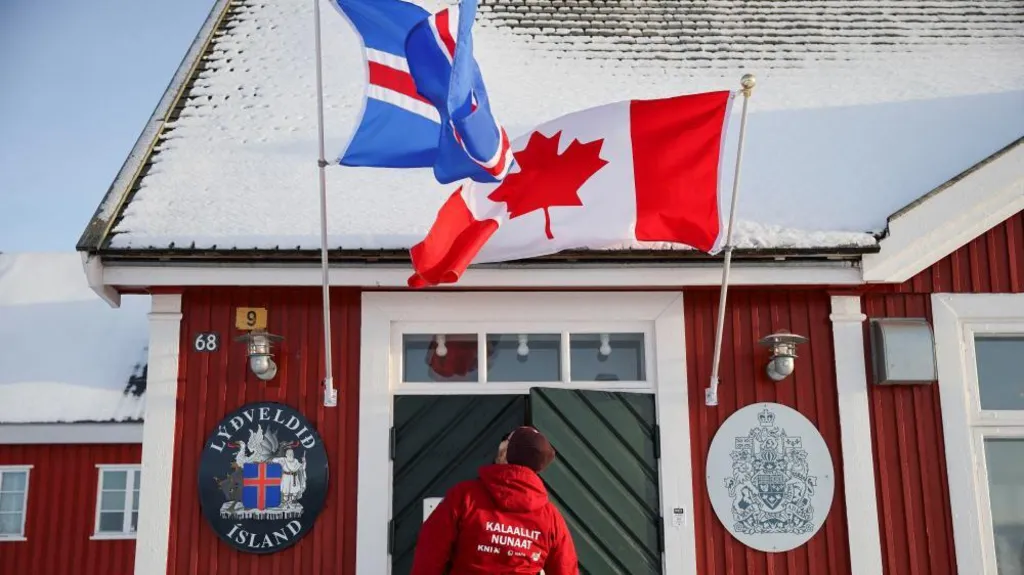 An image showing the front of the Canadian consulate building in Nuuk. It is a small building with a triangle roof and a double-door that is dark green with a white frame. On either side of the door are the official crest of Canada and the official crest of Iceland. Above the door are two big flags of Iceland and Canada.