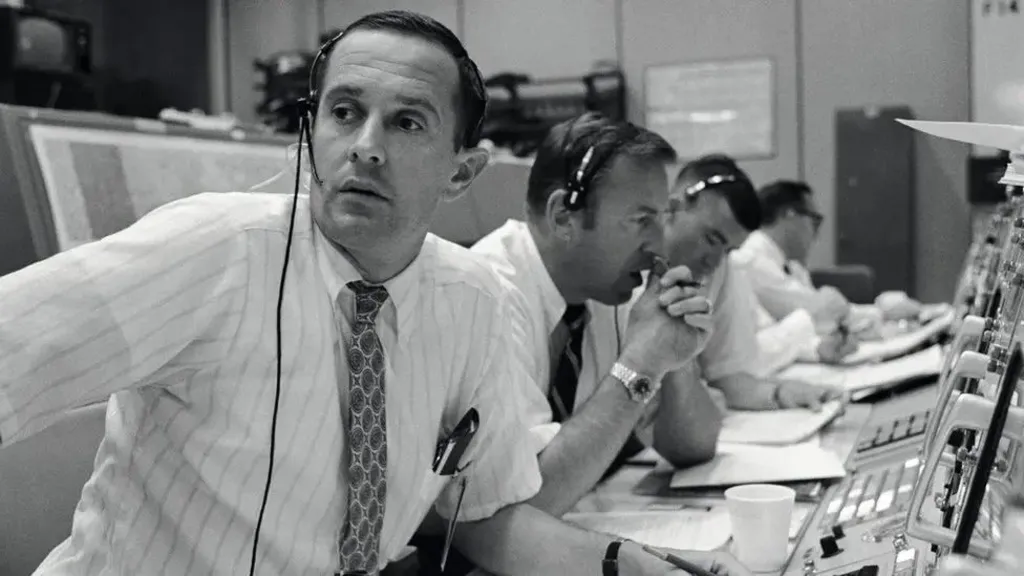 Black and white image of Charlie Duke, along with Jim Lovell and Fred Haise in Nasa Mission Control, during the Apollo 11 mission. They are all dressed in shirts and ties and wear headsets, as they sit in a long line in front of control panels and display screens.