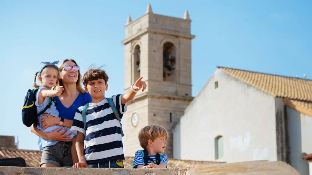 A woman wearing a tshirt, shorts and sunglasses holds a young girl wearing a backpack while two young boys stand in front of her looking and pointing into the distance. They're in front of a white bell tower in a European town.