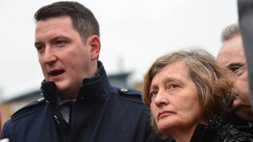 John Finucane is wearing a navy coat. He has brown hair. Geraldine Finucane is wearing a black coat. Other people are standing close to them. 