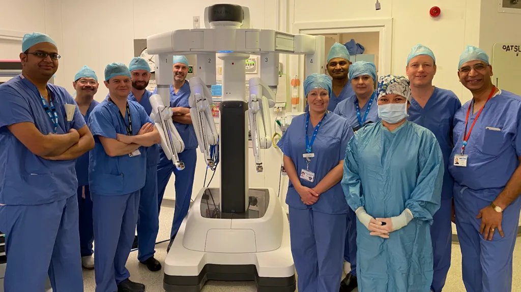 A group of people dressed in blue hospital scrubs, standing around a white piece of robotic equipment. They are standing in a light-coloured room, which appears to be a hospital setting.
