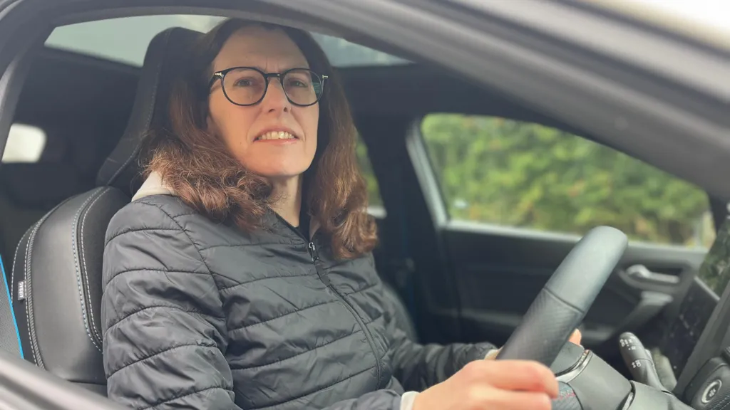 Image shows Caroline sat in her car. The photo is taken outside of the car and you can see the frame of the window. She is wearing a black puffer coat. She has black glasses on and is smiling at the camera. Her hair is brown and falls just beneath her shoulders. Her hands are holding the wheel. The car interior is black. 