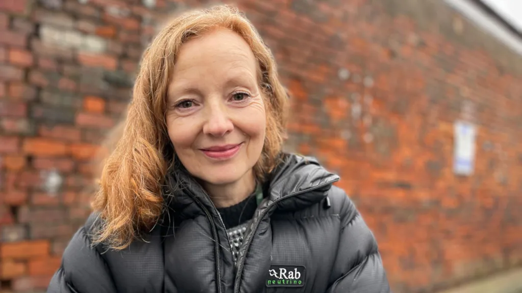 Sue McCutcheon smiles at the camera with her mouth closed. Her red hair is swept into a rough parting on her left. She is wearing a woolen jumper with a black/white/grey pattern and a black mountaineering coat over the top. She is sitting askew on a low wall with another adjacent red brick wall in the background