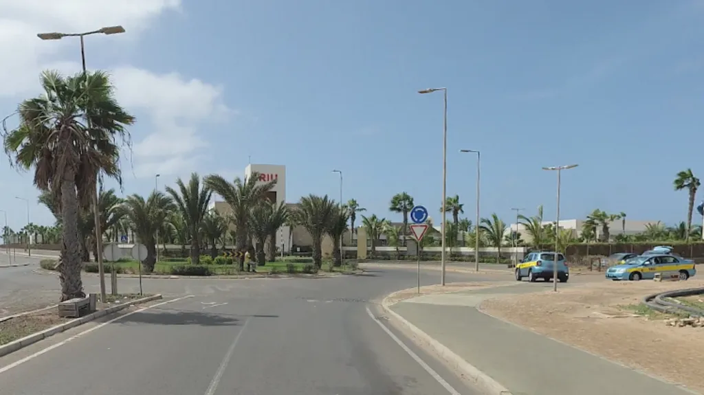 A street view of a hotel complex. There are palm trees and light blue taxis parked up on a roundabout outside of the hotel. In the background there is a white building with red letters saying 'RIU'. It appears to be a sunny day and the sky is blue.
