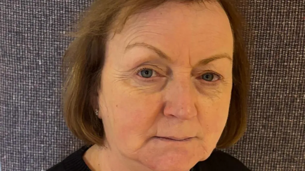 A close up shot of Mary Coyle who has short brown hair and is wearing a black top. She is standing in front of a grey fabric background.